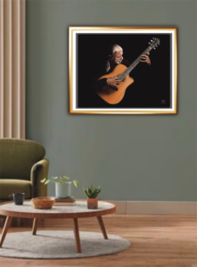 Musician in Concert. A man with silver hair in a ponytail, wearing a black jacket sits against a black background playing a Taylor Guitar, framed and displayed on a grey wall in a living room.